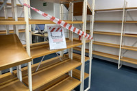 Leere Regale sind mit einen rot-weißen Band versehen. Auf einem Regalbrett steht ein Schild, das auf die Umbauarbeiten in der Bibliothek hinweist.