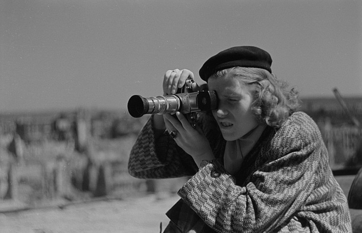 Frau in Mantel und Baskenmütze mit Kamera in der Hand, konzentriert fotografierend