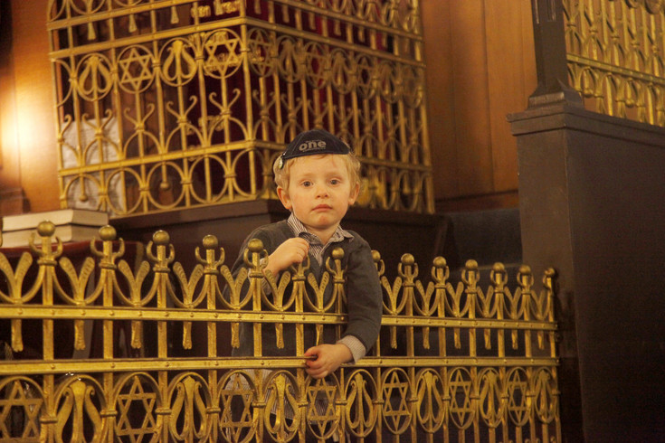 Ein kleiner blonder Junge stützt sich auf einen goldfarbenen kunstvoll verzierten Gitterzaun innerhalb einer Synagoge.