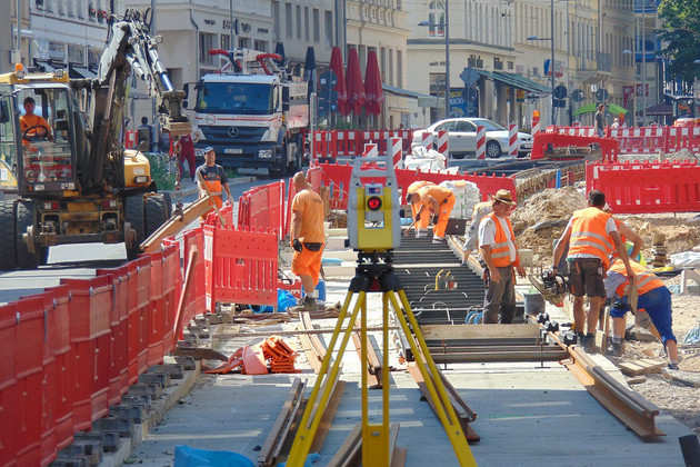 Baustelle auf der Karl-Liebknecht-Straße mit Bauzäunen, Baumaschinen und Arbeitern. Ein Gleis wird auf einem Betonbett verlegt.