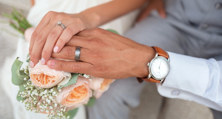 Die Hände einer Frau und eines Mannes liegen auf einem Blumenstrauß. Man erkennt im Hintergrund, dass die Frau ein weißes Kleid und der Mann einen grauen Anzug trägt. Beide tragen Eheringe.