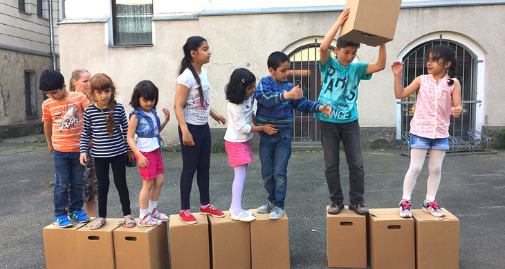 Kinder unterschiedlicher Herkunft stehen in einer Reihe auf Kartons und reichen siche einen Karton weiter.