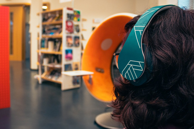 Hinterkopf einer Frau mit Brille und Over-Ear-Kopfhörer. Im Hintergrund steht ein orange-farbener Sonic Chair und Bücherregale.