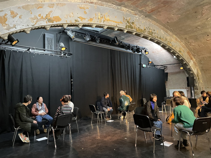 In einem Theaterraum sind auf einer Bühne vor einem schwarzem Vorhang Stuhlkreise aufgebaut. Es stehen insgesamt vier Kreise mit jeweils vier Stühlen auf der Bühne. Auf den Stühlen sitzen drei bis vier Personen unterschiedlicher Nationen, die in Gesprächen vertieft sind.