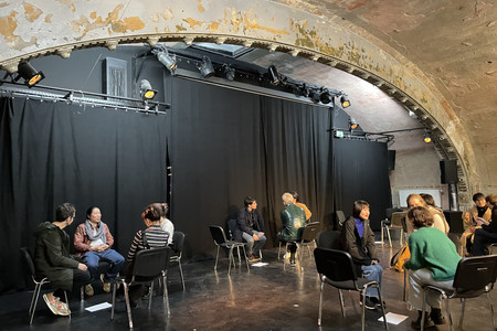 In einem Theaterraum sind auf einer Bühne vor einem schwarzem Vorhang Stuhlkreise aufgebaut. Es stehen insgesamt vier Kreise mit jeweils vier Stühlen auf der Bühne. Auf den Stühlen sitzen drei bis vier Personen unterschiedlicher Nationen, die in Gesprächen vertieft sind.