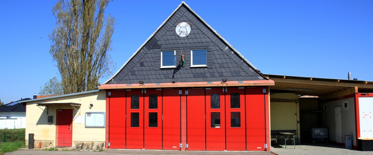 Das Gebäude der Freiwillige Feuerwehr Knautnaundorf mit zwei großen, roten Hallentoren nebeneinander. Darüber ein Spitzdach. Die Giebelseite ist mit Schiefer gedeckt, besitzt zwei Fenster und wird durch ein Feuerwehrsymbol geziert.