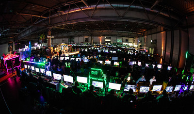 In einer abgedunkelten Halle sind viele Computermonitore nebeneinander. Daran spielen viele Menschen Computerspiele.