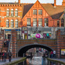 Kanal fließt in Birmingham unter einer roten Backsteinbrücke, welche oberhalb mit roten Backsteingebäuden bebaut ist. Diese Gebäude beherbergen gastronomische Einrichtungen.