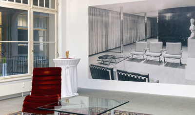 roter Sessel steht neben einem Glastisch und vor einer Wand mit einer historischen schwarz-weiß Fotografie desselben Raumes