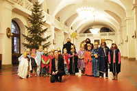 Eine Frau hockt vor 14 Kindern in Kostümen mit Kronen vor einem leuchtenden Weihnachtsbaum in einer mondänen Säulenhalle