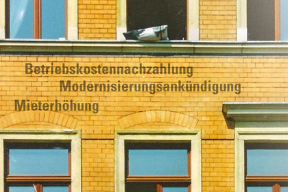 Kleiner Ausschnitt einer Altbaufassade. Zwischen zwei Stockwerken stehen untereinander sie Worte "Betriebskostennachzahlung", "Modernisierungsankündigung" und "Mieterhöhung".