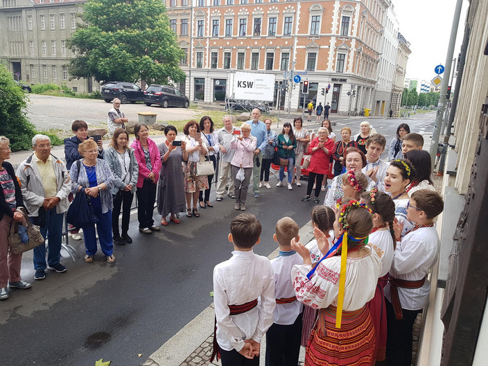 Viele Menschen stehen auf der Straße und hören einem Kinderchor zu. Die Kinder tragen traditionelle ukrainische Kleidung, die Mädchen im Chor bunte Blumenkränze auf dem Kopf und Schleifen im Haar.