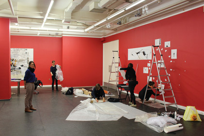 Bilder werden mithilfe einer Leiter an rote Wände in einer Galerie aufgehangen.
