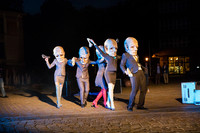 Fünf Schauspieler und Schauspielerinnen mit Anzug beziehungsweise Kostüm bekleidet tanzen auf einem städtischen Platz. Alle tragen als Maske einen überdimensionalen Kopf eines alten, faltigen Mannes mit Glatze.