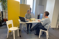 In einem abgetrennten Bereich in der Bibliothek Reudnitz sitzen zwei Frauen am Tisch und unterhalten sich. Auf dem Tisch sind ein Laptop, Bücher, Papier und Stifte zu sehen.