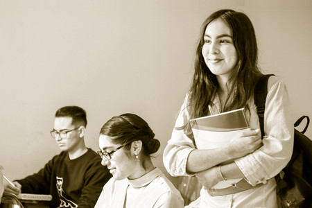 Vier Jugendliche sitzen in einem Klassenraum, ein Mädchen mit Büchern steht in der Mitte.