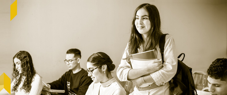 Vier Jugendliche sitzen in einem Klassenraum, ein Mädchen mit Büchern steht in der Mitte.