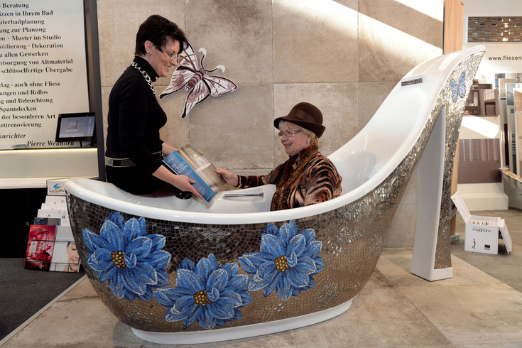 Eine alte Dame mit Hut sitzt Probe in einer freistehenden Badewanne auf einer Messeausstellung.