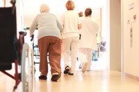 Eine alte Frau geht am Rollator einen Gang entlang, vor ihr zwei Pflegekräfte in weißer Kleidung, vorn am Bildrand ein Rollstuhl