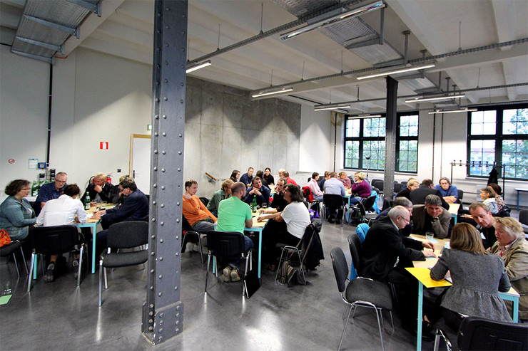 Blick in eine Halle, in der ca. 50 Menschen an verschiedenen Tischen sitzen.