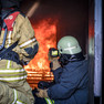 Zwei Feuerwehrmänner sitzen in Feuerschutzkleidung in einem Container für Brandversuche vor einem Feuer. Einer von ihnen beobachtet das Feuer durch eine Wärmebildkamera