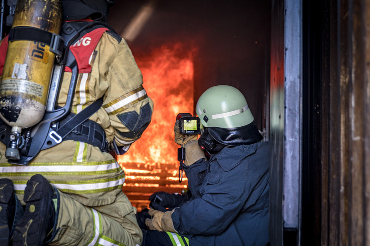 Zwei Feuerwehrmänner sitzen in Feuerschutzkleidung in einem Container für Brandversuche vor einem Feuer. Einer von ihnen beobachtet das Feuer durch eine Wärmebildkamera