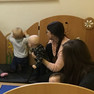 Zu sehen sind mehrere Kleinkinder und zwei Mütter, die mit den Kindern vor einer Spielwand aus Holz sitzen. Zwei der Kinder sitzen bei den Müttern auf dem Schoß, die anderen beiden Kindern beschäftigen sich mit der Spielwand aus Holz. 