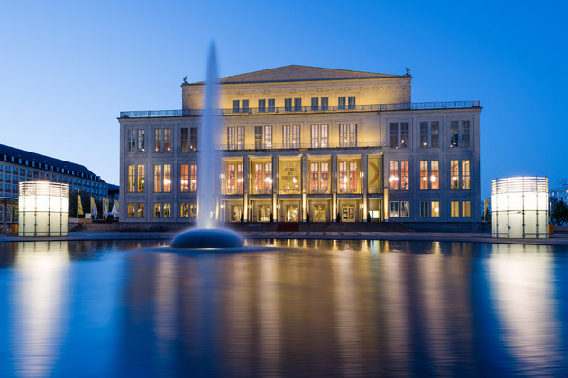 Außenansicht der Oper Leipzig zur blauen Stunde mit einer flachen Wasserfläche und einer Wasserfontäne davor.