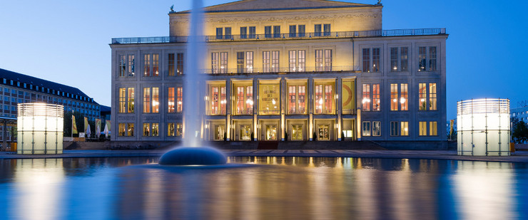 Außenansicht der Oper Leipzig zur blauen Stunde mit einer flachen Wasserfläche und einer Wasserfontäne davor.