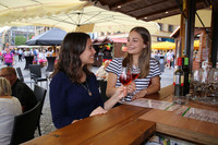 Zwei junge Frauen stoßen an einem hölzernen Marktstand mit Weingläsern an.