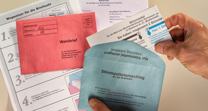 eine Hand hält blauen Wahlzettelumschlag und weitere Unterlagen zur Briefwahl liegen auf dem Tisch
