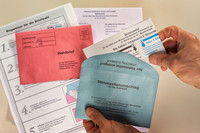 eine Hand hält blauen Wahlzettelumschlag und weitere Unterlagen zur Briefwahl liegen auf dem Tisch