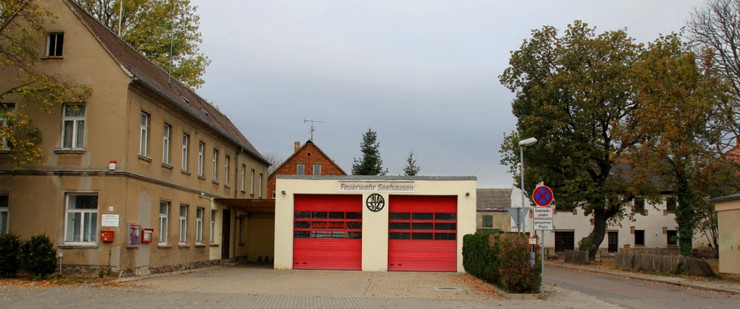 In der Mitte eine Garage mit zwei roten Rolltoren. Dahinter ein Feuerwehrauto. Zwischen den beiden Toren ist ein schwarzes Feuerwehrsymbol und darüber steht in großen, roten Buchstaben "Feuerwehr Seehausen". Links neben der Halle ein zweietagiges Gebäude, davor ein gepflasterter Vorplatz.