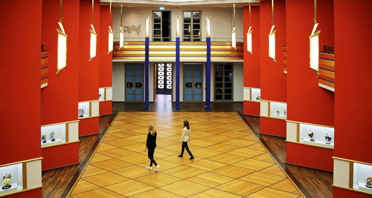 Eine hohe Halle mit schräg zueinander stehenden roten Säulen und Parkettboden. Zwei Frauen laufen durch die Halle.