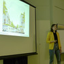 Frau in gelben Blazer hält eine Präsentation, sichtbar ist auch eine Präsentationswand.