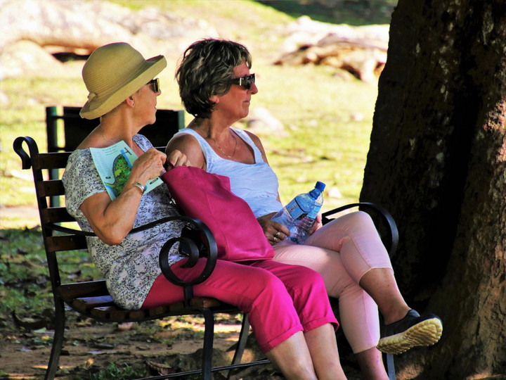 Zwei Seniorinnen sitzen im Schatten auf einer Bank. Eine trägt einen Sonnenhut, die andere hat zwei Wasserflaschen in der Hand