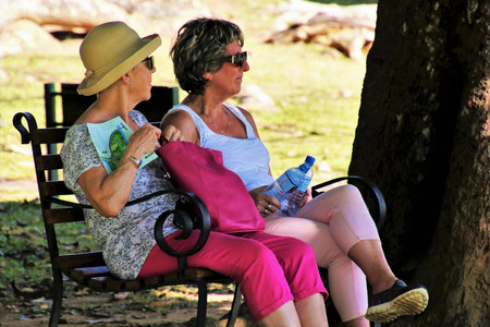 Zwei Seniorinnen sitzen im Schatten auf einer Bank. Eine trägt einen Sonnenhut, die andere hat zwei Wasserflaschen in der Hand