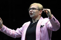 Mann mit blondgefärbten Haaren, bei denen der dunkle Ansatz zu sehen ist, steht in einem rosa Jackett und mit großer markanter Brille gestikulierend auf der Bühne.