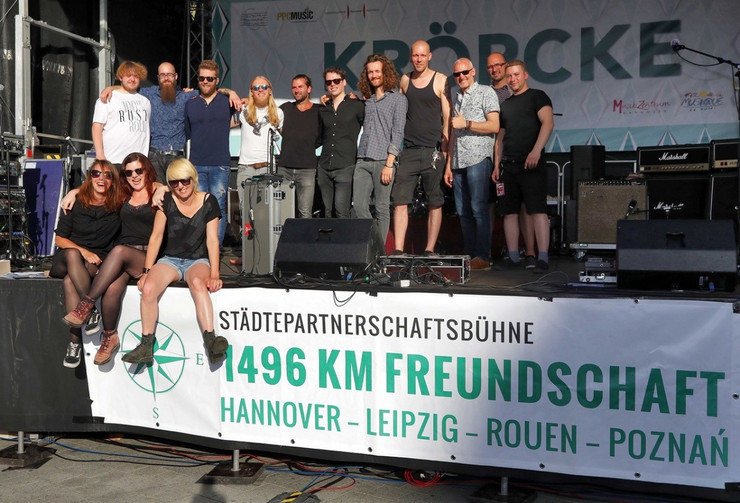 Bühne mit dem Banner "Städtepartnerschaftsbühne", auf der 14 Akteure für ein Gruppenbild posieren, darunter die drei Musiker der Leipziger Band Lizard Pool.