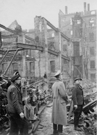 Drei Männer auf einem in Trümmern liegenden Arial mit zerstörten Gebäuden im Hintergrund