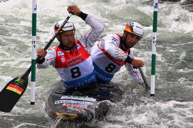 Zwei Kanusportler sitzen in einem Kanu und paddeln mit je einem Paddel in den stür,ischen Wellen durch zwei Zielstangen