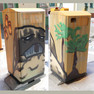 Abfallbehälter mit Graffiti-Motiv, eine Figur und zwei Bäume