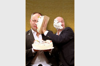 Zwei Männer in Anzügen. Einer hält eine Torte in den Händen, der andere wirft sie ihm ins Gesicht.