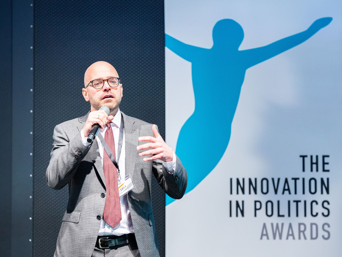 Ein Mann steht vor einem Roll up auf dem The Innovation in Politics Award steht und spricht in ein Mikrofon.