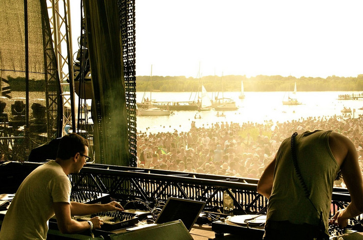 Zwei DJs an ihren Mischpulten. Im Hintergrund das Publikum im Sonnenuntergang und der Cospudener See mit Segelbooten.