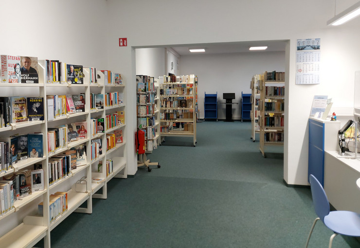 Links viele Bücherregale, in der Mitte der lange Gang mit Blick durch die Bibliothek und rechts die Verbuchungstheke mit einen blauen Stuhl.