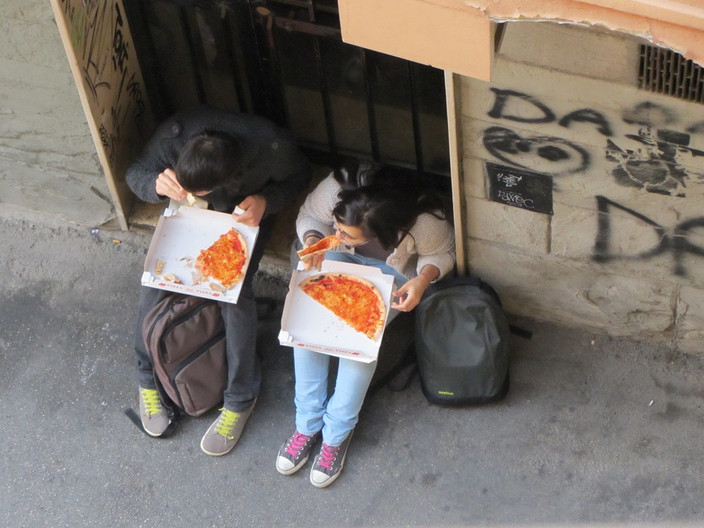Blick vom Balkon auf zwei in einer Gasse sitzende Personen, die Pizza essen