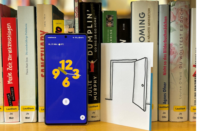 Handy mit Uhrzeitanzeige neben Blatt Papier auf dem eine geöffnete Tür gezeichnet ist. Beides steht in einem Bücherregal vor Büchern.