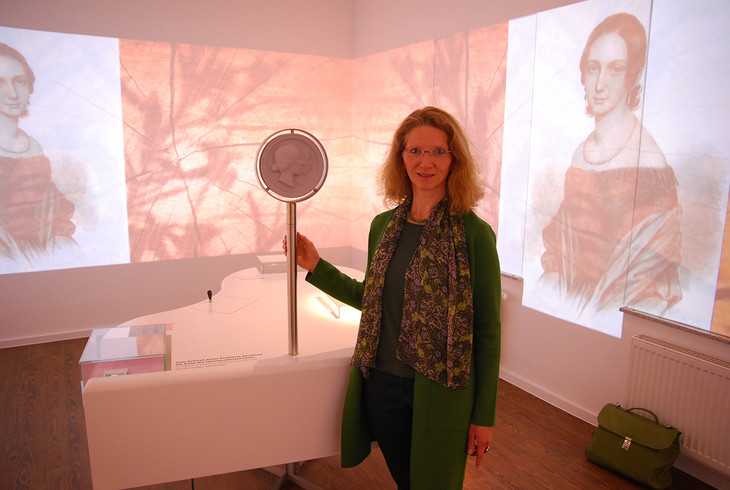 Hörfilmproduzentin Magdalene Melchers steht in einem Ausstellungsraum vor einem weißen Flügel. An die Zimmerwände sind Bilder von Clara Schumann projiziert.
