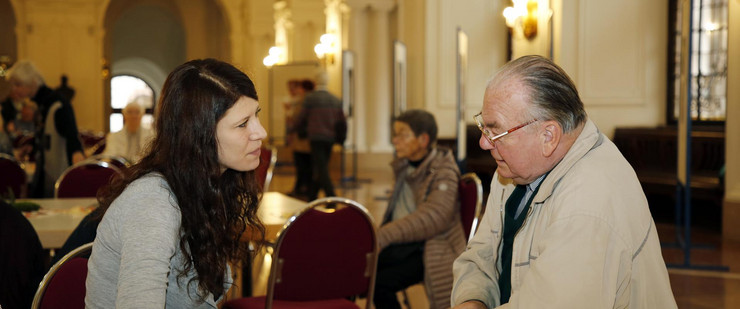 Eine junge Frau und ein älterer Herr sitzen an einem Tisch und diskutieren miteinander.
